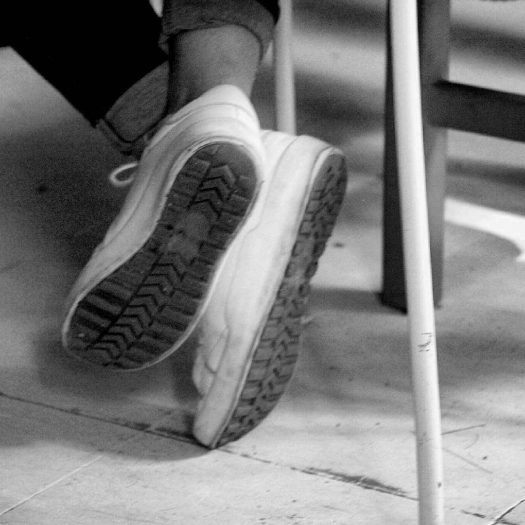 Black and white photograph showing the soles of white sneakers resting on a tiled floor, with a metal pole visible in the frame.